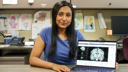 Ria Patel sitting and smiling at a table with an x-ray of a brain on a laptop screen next to her.