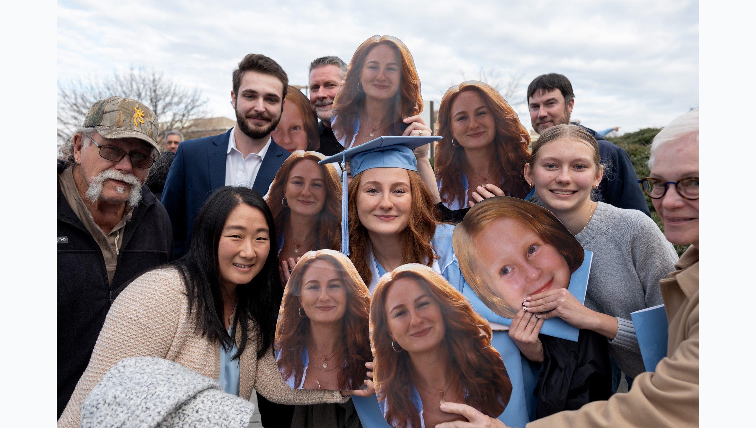 A female graduate surrounded by family members and/or friends following U.N.C. Chapel Hill's Winter Commencement. Those around her are holding up face cutout signs of the graduate.