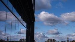 Long-distance shot of the Morehead-Patterson Bell Tower on the campus of U.N.C Chapel Hill on a sunny day. Clouds are seen in the background, as is a reflection of the scene, bouncing off the windows of a building.